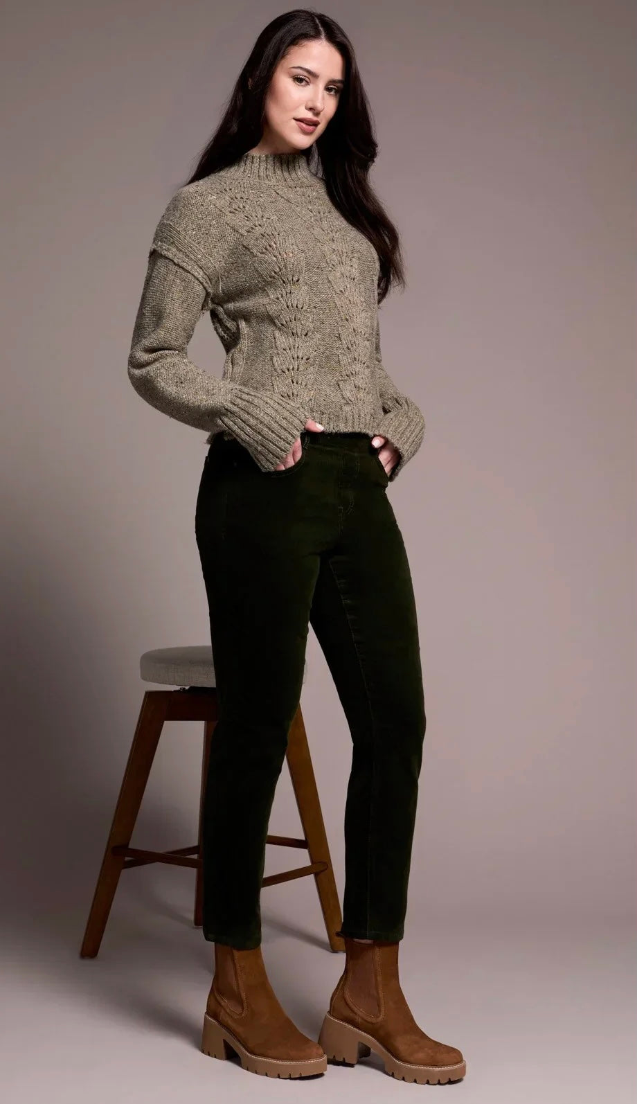 A woman modeling a textured taupe cable-knit mock-neck sweater and dark green cropped pants, standing next to a wooden stool against a neutral studio backdrop.