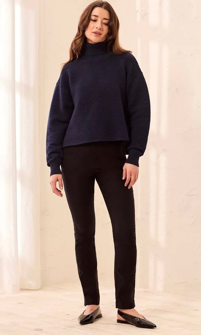 A woman modeling a dark navy ribbed knit mock neck sweater, paired with black pants and black flats, standing in a brightly lit cream-colored room.