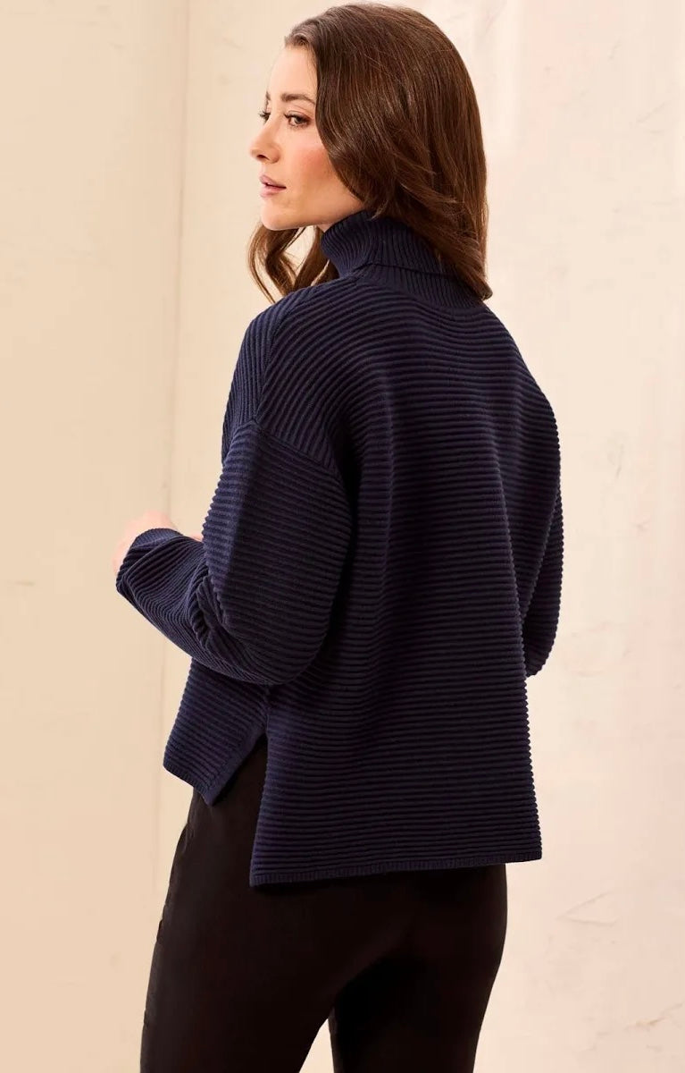A woman wearing a dark navy ribbed knit mock neck sweater, shown from a side-back angle against a neutral beige wall.