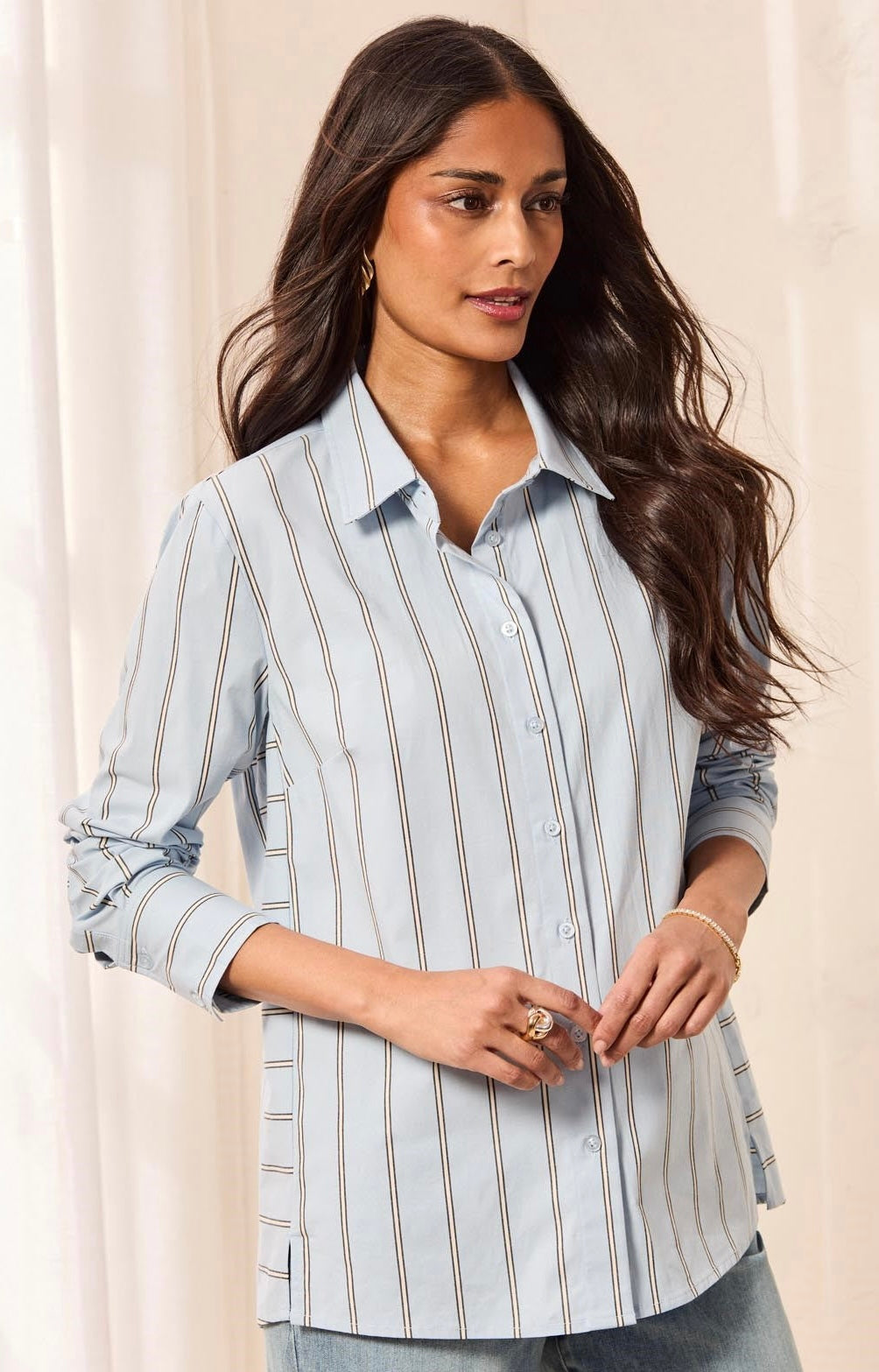 A woman wearing a light blue and white vertically striped button-up shirt with rolled-up sleeves, paired with blue jeans, set against a soft beige curtain background.