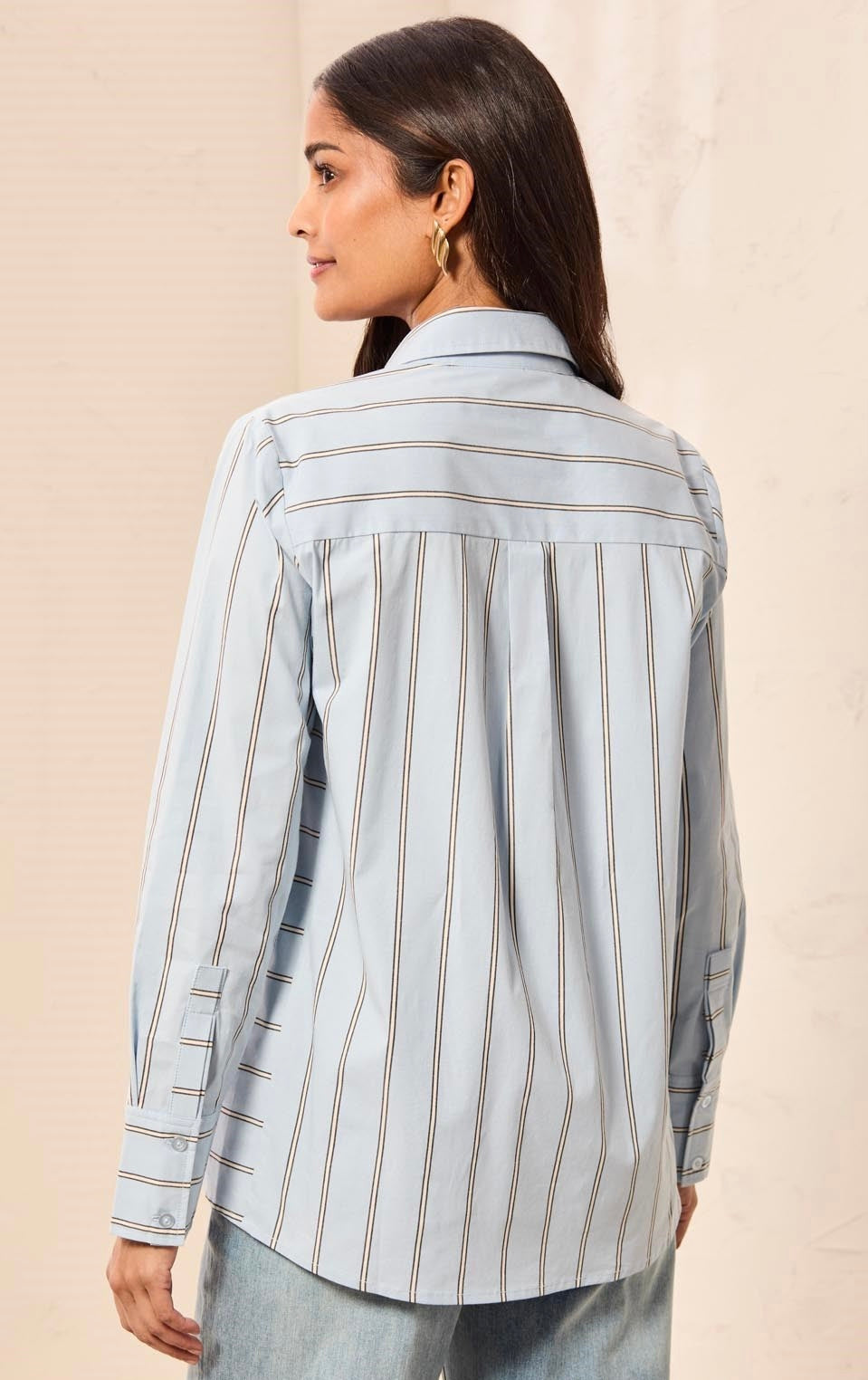 A woman shown from the back wearing a light blue striped button-up shirt with thin brown vertical lines, paired with light-wash jeans, against a neutral beige background.