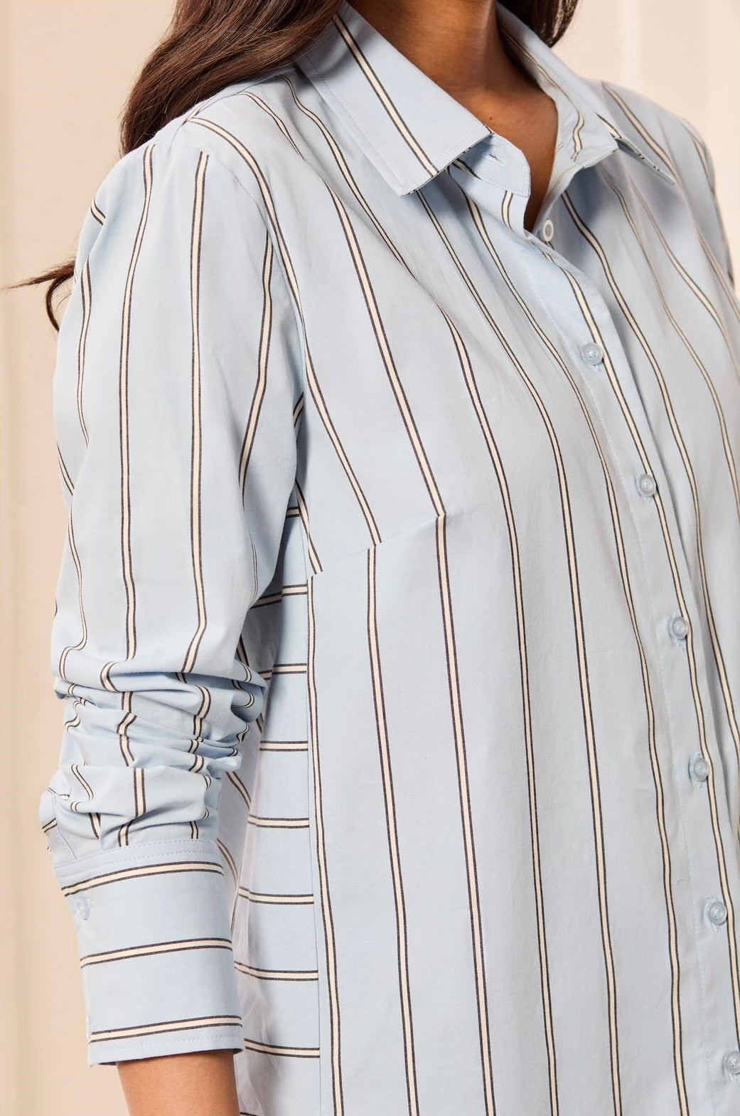 A close-up of a woman wearing a light blue button-up shirt with vertical brown stripes, showing the collar, buttoned front, and rolled-up sleeve.