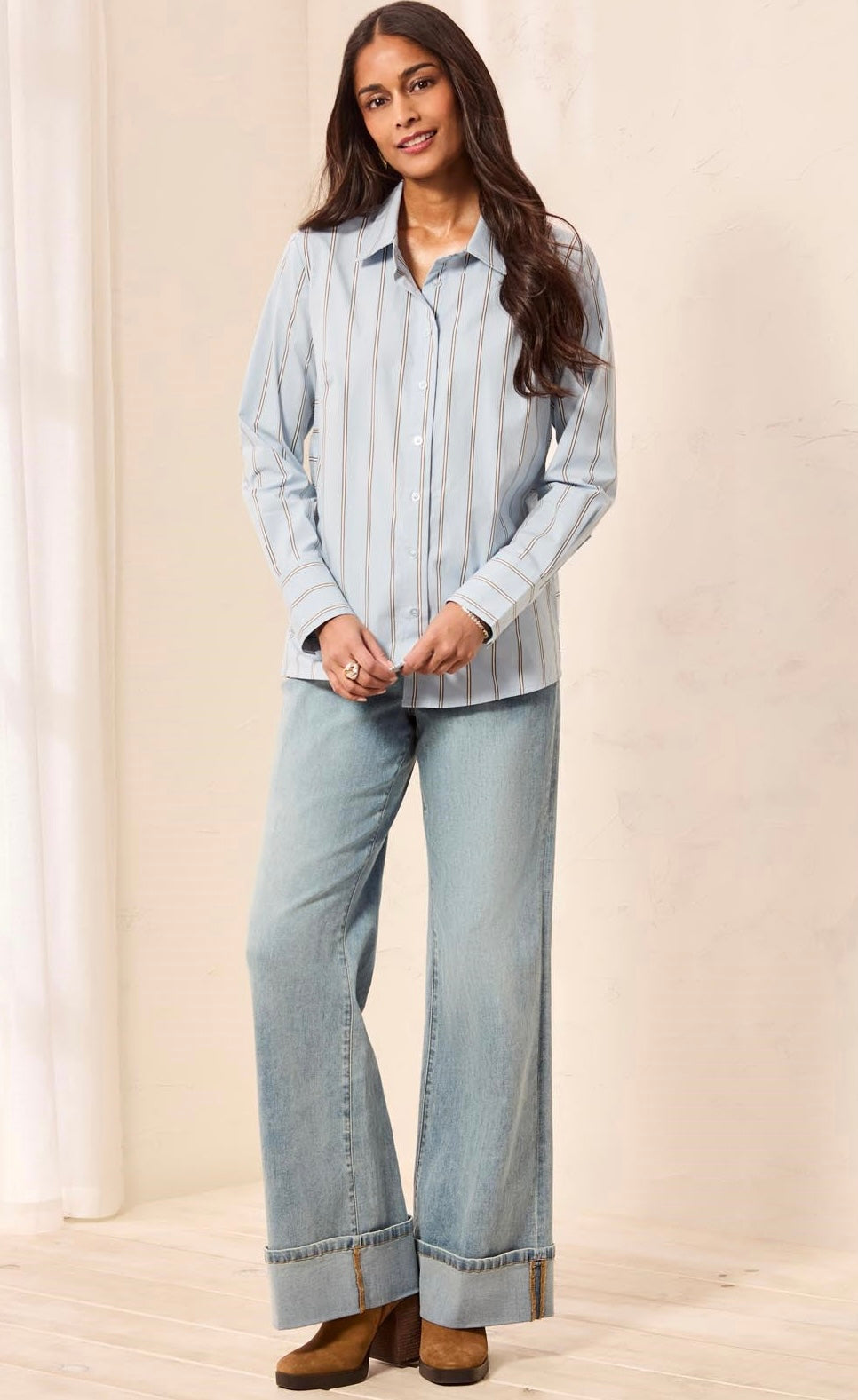 A woman modeling a light blue and white vertically striped long-sleeve button-up shirt paired with wide-leg light blue jeans and brown ankle boots, standing against a warm beige studio backdrop.