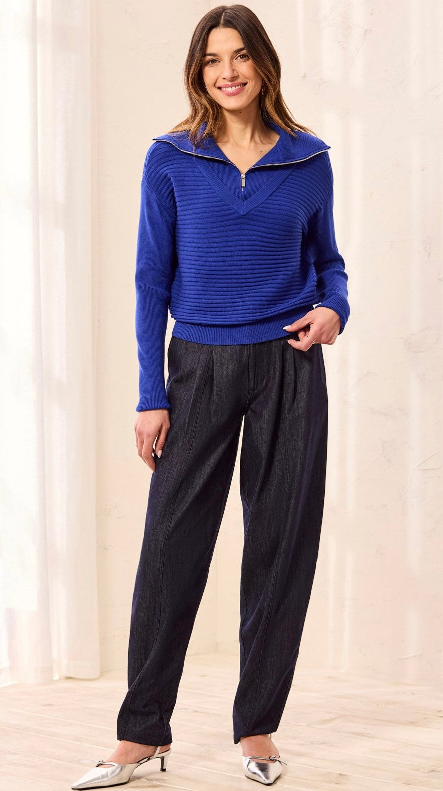 A woman modeling a royal blue ribbed knit half-zip sweater with a wide collar and dark charcoal pleated trousers, standing in a bright indoor setting with light curtains and a wooden floor.