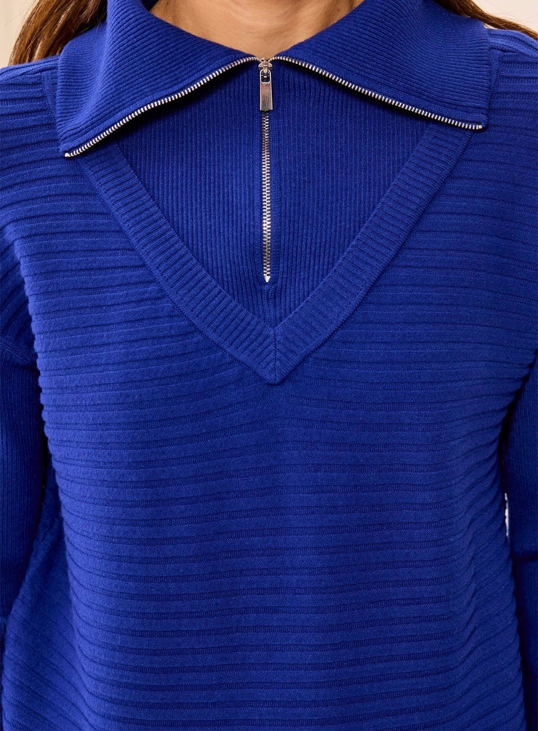 A close-up of a person wearing a royal blue ribbed knit sweater with a V-neck and a short front zipper.