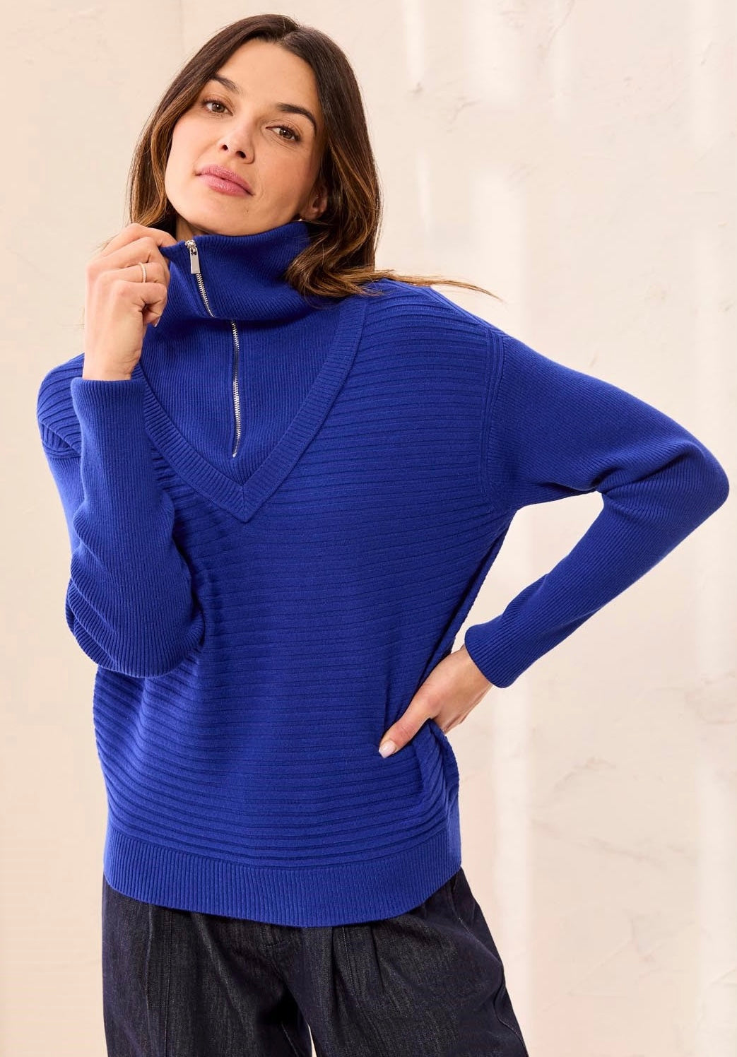A woman wearing a vibrant royal blue ribbed knit sweater with a half-zip mock neck, paired with dark gray pants, posing against a light beige wall.
