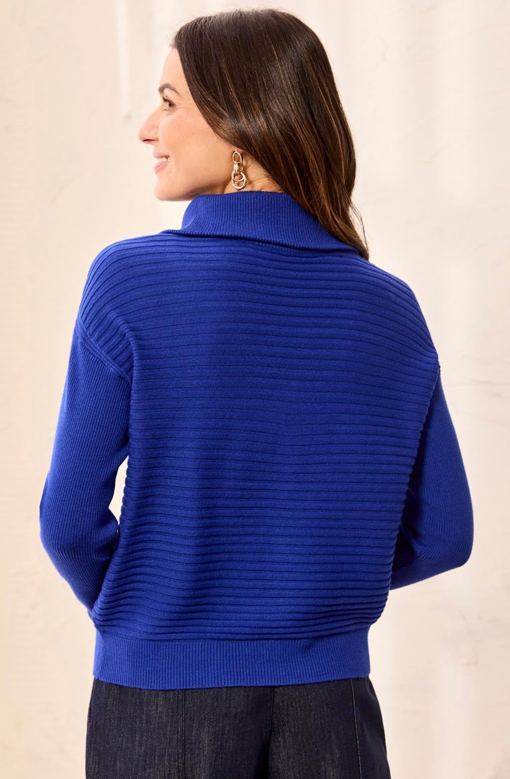 A woman shown from the back wearing a bright royal blue ribbed knit mock neck sweater, paired with dark gray pants, against a neutral beige background.
