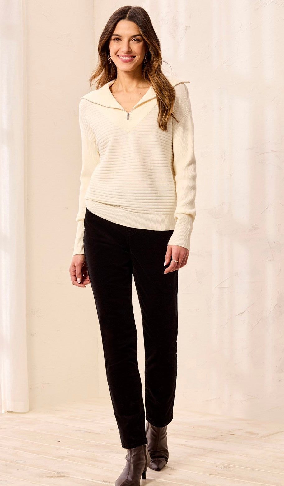 A woman modeling a cream ribbed half-zip mock-neck sweater and black pants against a light beige curtain backdrop.