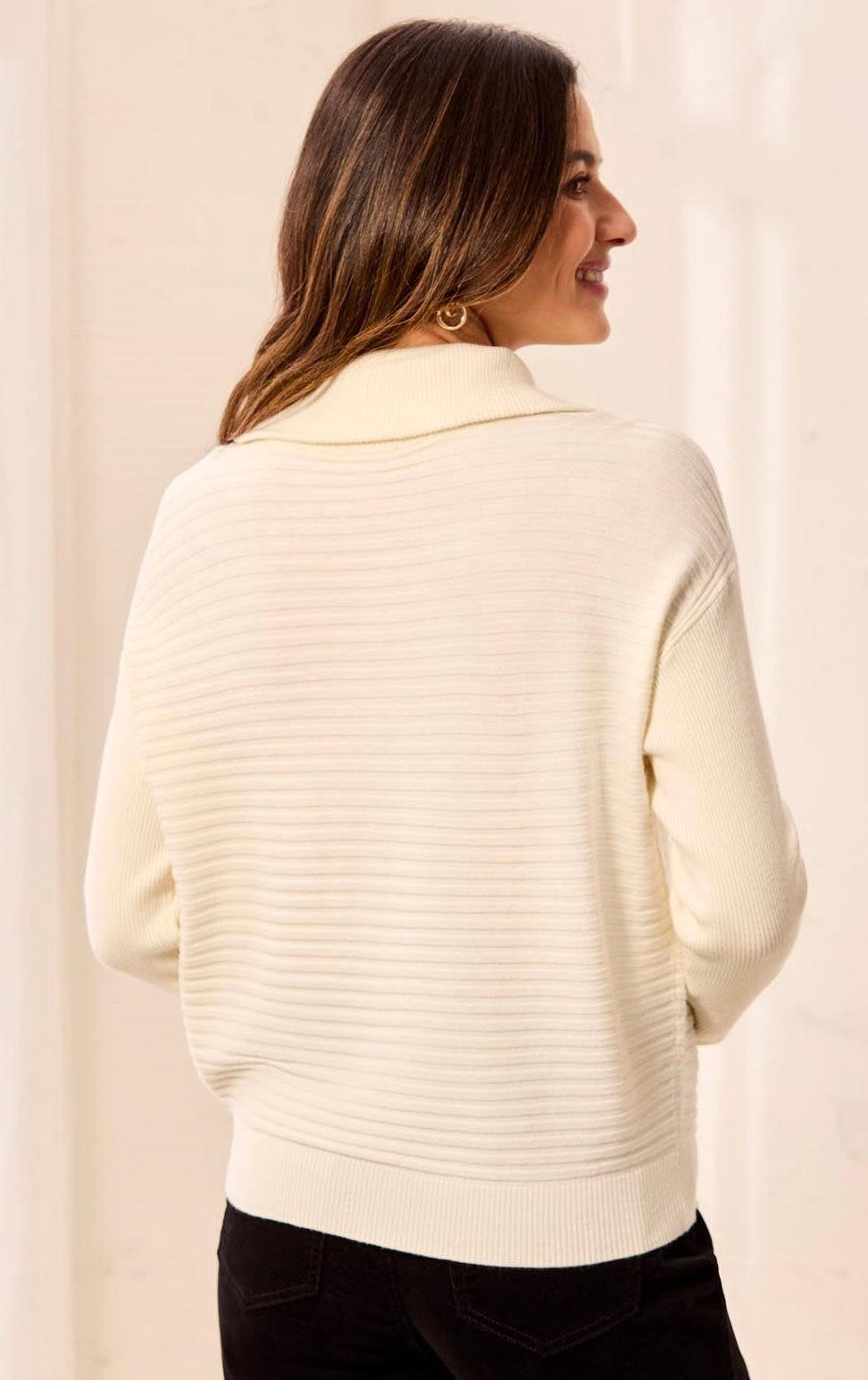 A woman shown from the back wearing a cream ribbed knit mock neck sweater and black pants, standing against a soft beige background.