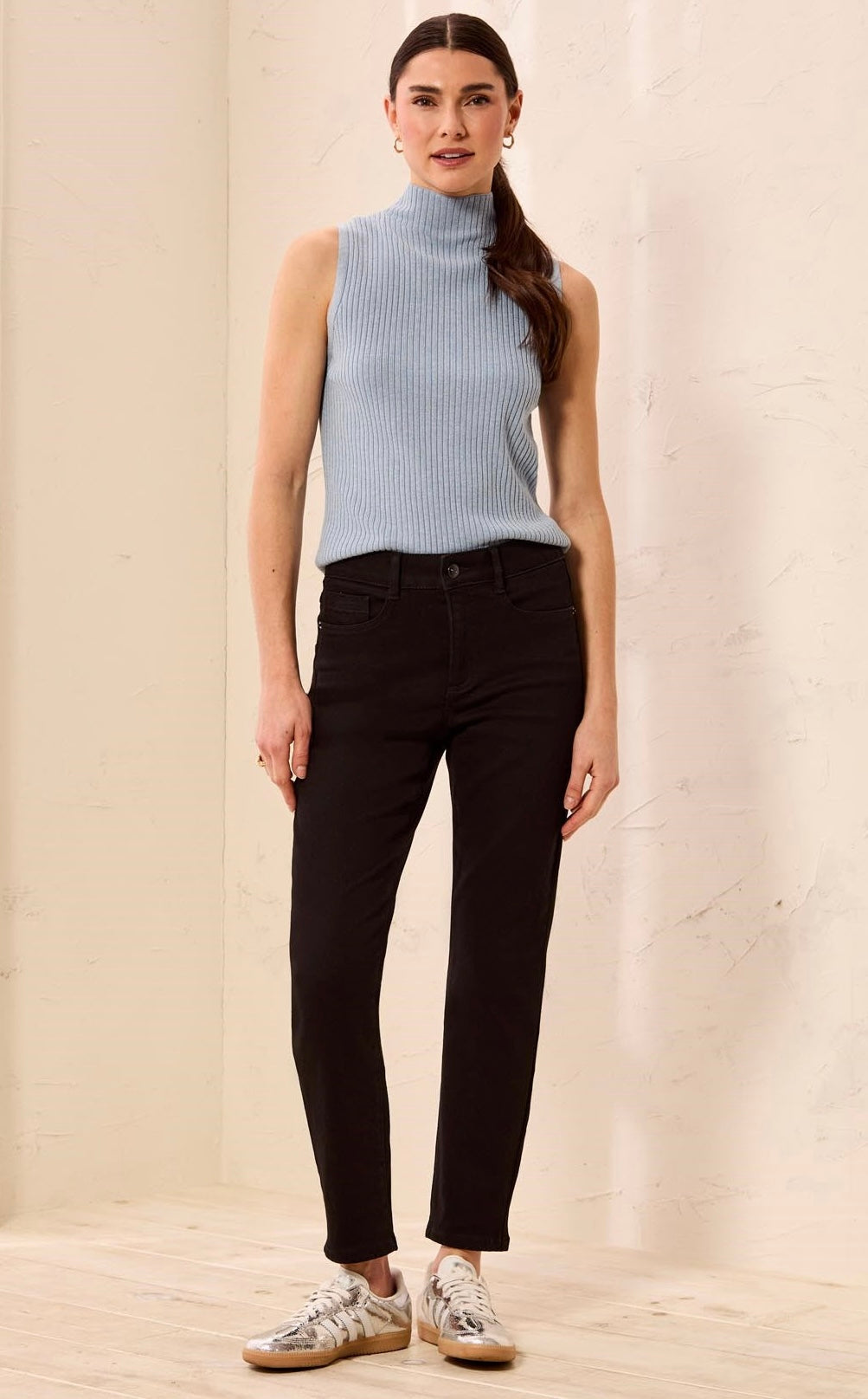 A woman modeling a sleeveless light blue ribbed turtleneck sweater vest paired with black skinny jeans and white sneakers, standing against a neutral beige textured wall.