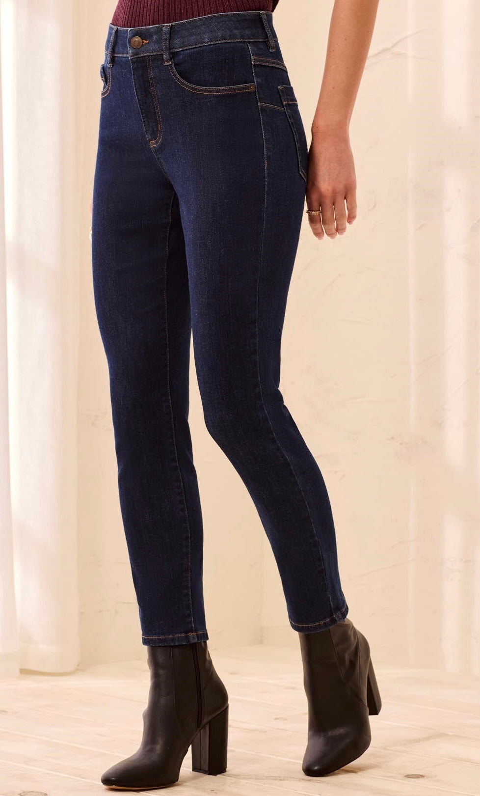 A person wearing dark blue skinny jeans with black ankle boots, shown from the waist down in a softly lit indoor setting.