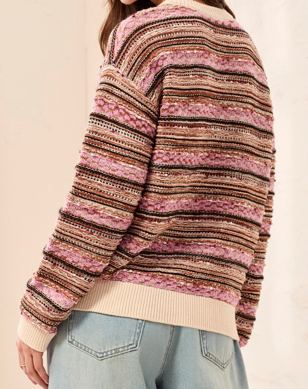 A person wearing a striped knit sweater with horizontal bands of pink, beige, brown, and black, paired with light blue jeans, shown from a side-back angle against a plain light background.