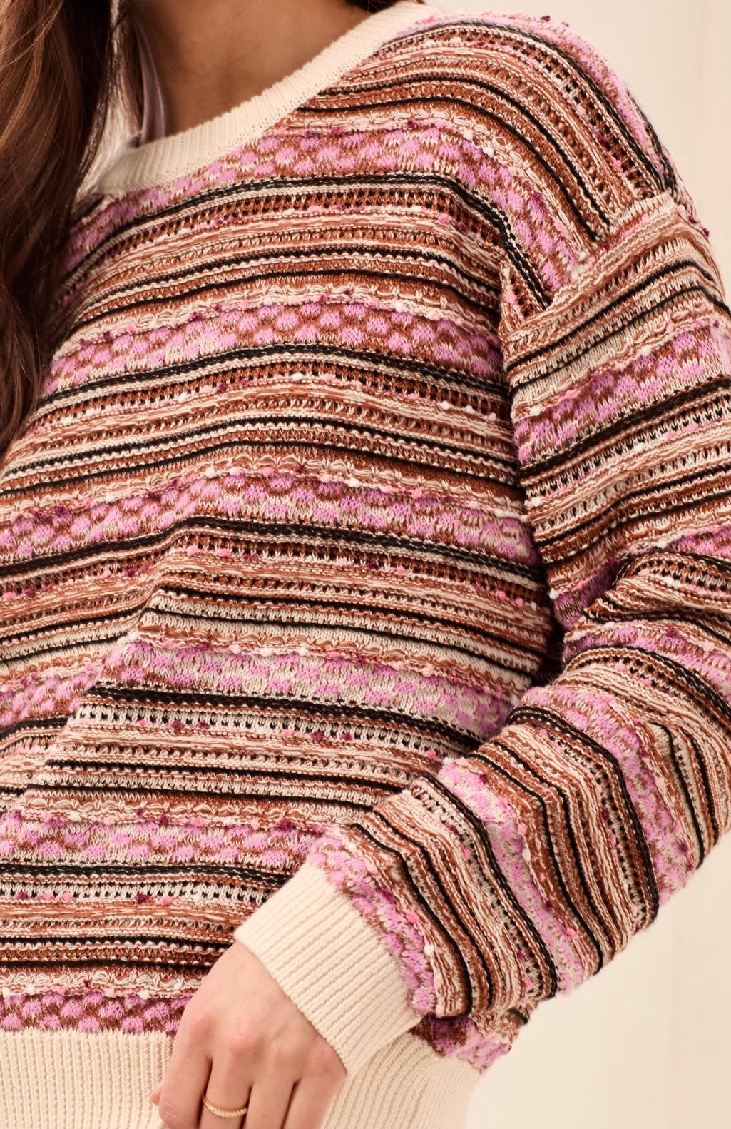 A close-up of a person wearing a textured knit sweater with horizontal stripes in pink, brown, beige, and black, featuring ribbed cream cuffs and hem.
