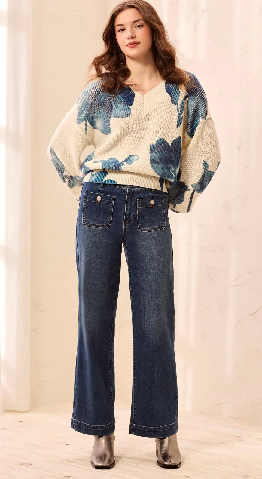 A woman modeling a cream knit sweater with blue floral patterns and wide-leg dark blue jeans, standing in a softly lit indoor setting.