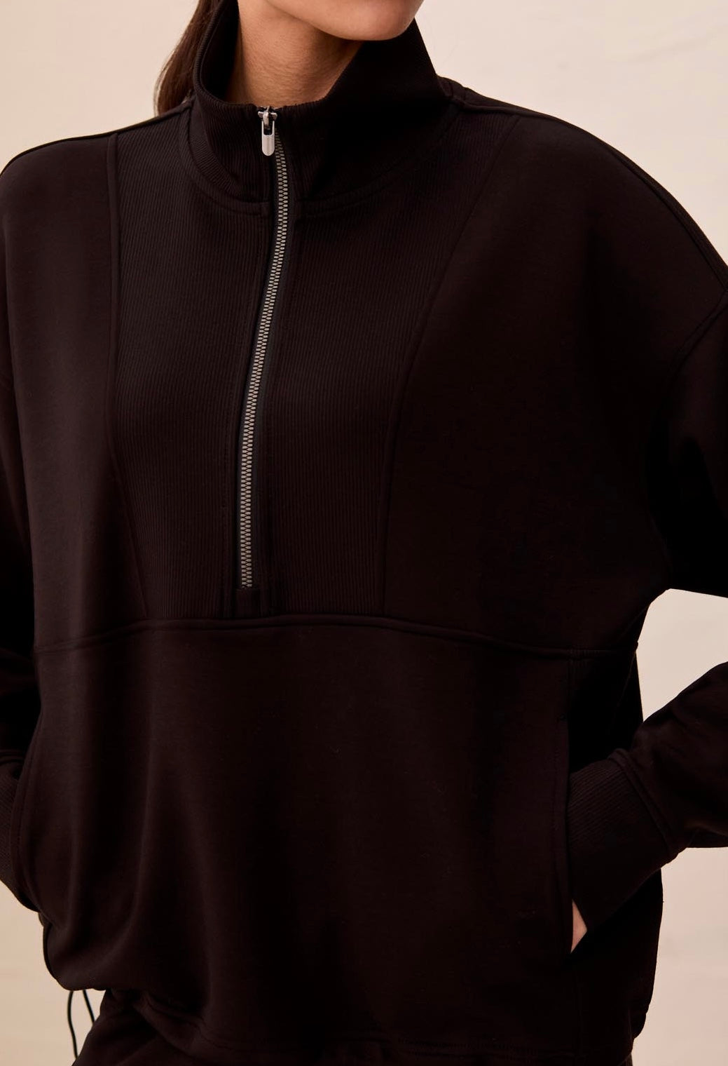 A close-up of a person wearing a dark brown or very deep black quarter-zip pullover sweatshirt with a stand collar and side pockets, against a plain light beige background.