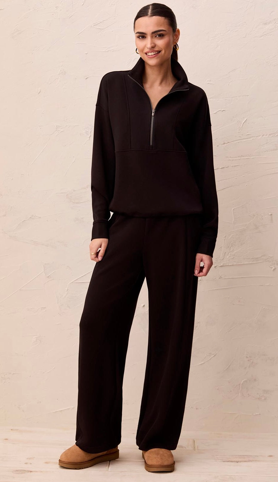 A woman modeling a matching dark brown lounge set consisting of a long-sleeve half-zip pullover and wide-leg pants, paired with tan suede slippers, against a textured beige wall.