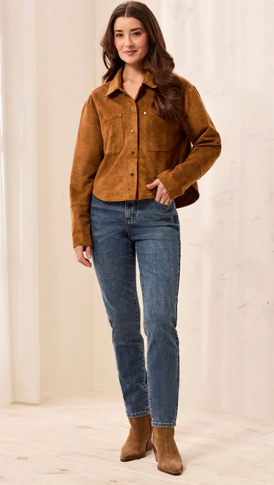 A woman modeling a cropped brown suede button-up shirt jacket, paired with blue jeans and brown suede ankle boots, standing in a bright, neutral indoor setting.