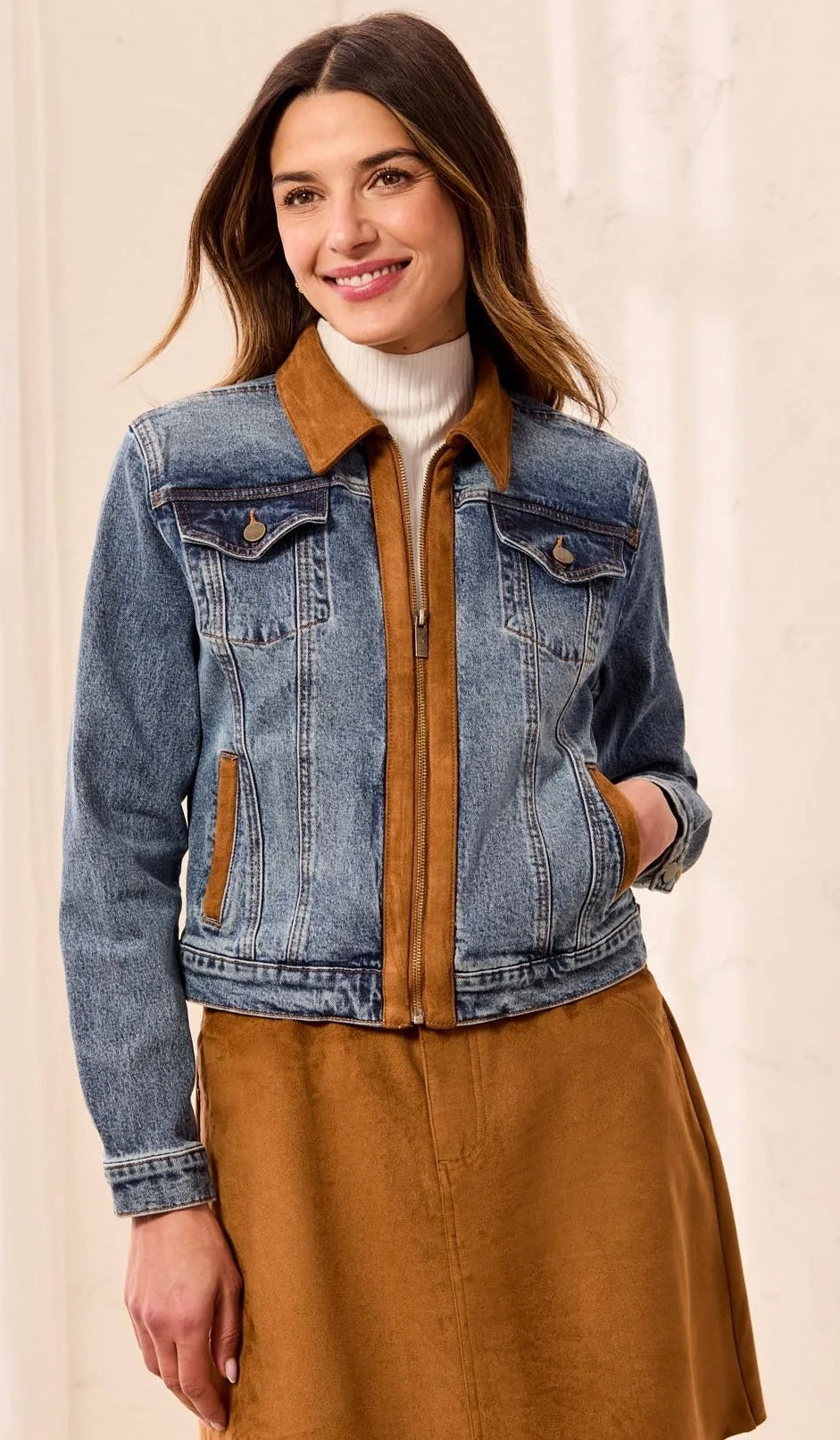 A woman wearing a blue denim jacket with a brown suede-like collar and front placket, layered over a white turtleneck and brown skirt, against a light beige background.