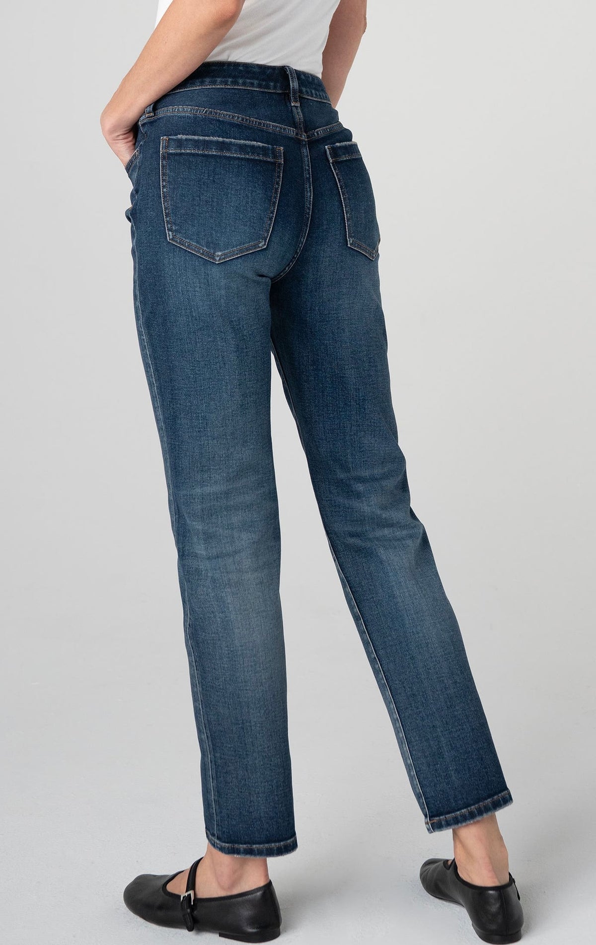 Rear view of a person wearing dark blue straight-leg jeans with a white top and black flats, standing against a light gray studio background.