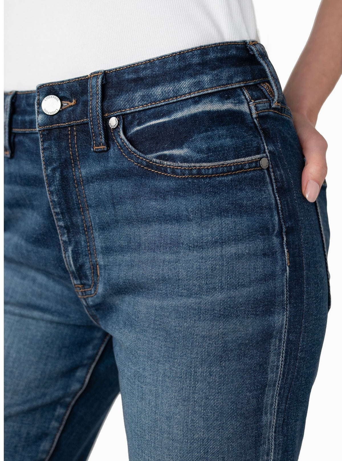 Close-up of a person wearing dark blue denim jeans with a white top, showing the front pocket, button, and hand in the side pocket against a plain white background.