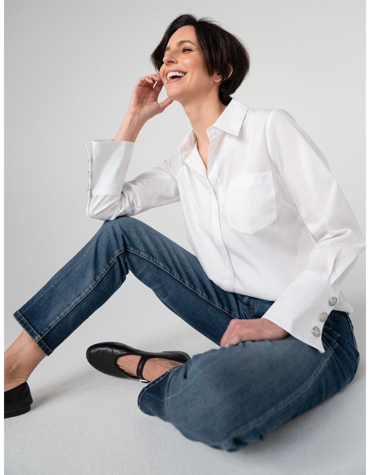 A woman sitting on a light gray floor wearing a white long-sleeve button-up shirt, blue jeans, and black Mary Jane shoes.