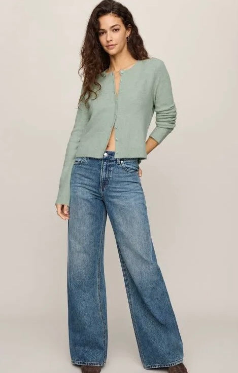 A woman modeling a sage green cropped cardigan with a split neckline over a matching top, paired with high-waisted, wide-leg blue jeans, against a solid light beige studio background.