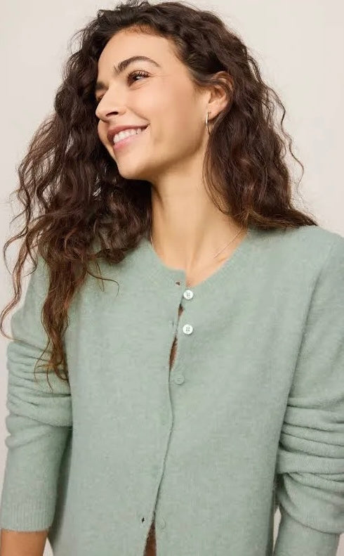 A woman with long, wavy brown hair smiling and looking to the side, wearing a sage green knit cardigan with a buttoned neckline over a brown top, set against a plain light beige background.