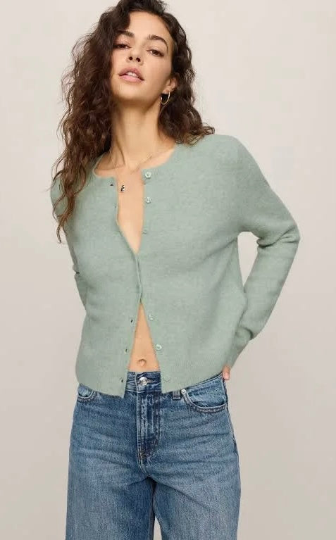 A woman wearing a sage green knit cardigan with silver buttons and blue jeans, posing against a neutral light beige background.