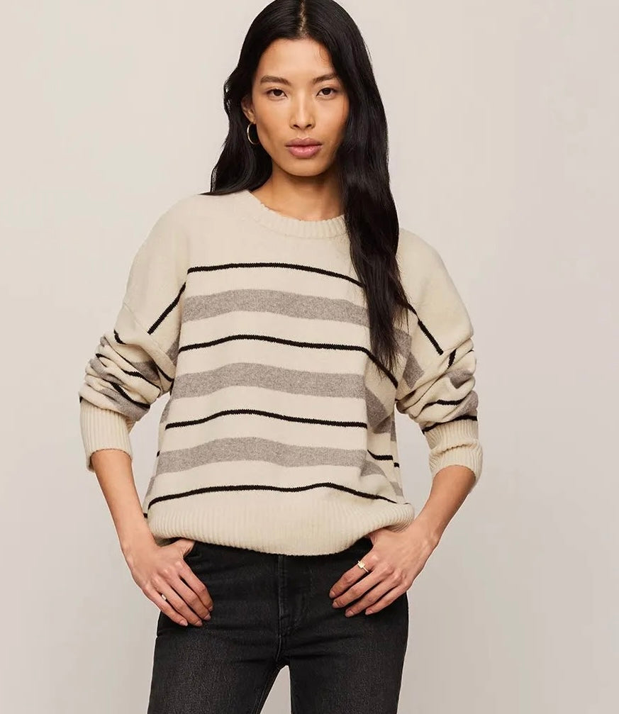 A woman modeling a cream-colored knit sweater with horizontal black and gray stripes, paired with dark jeans, against a plain light beige background.
