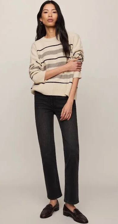 A woman modeling a cream knit sweater with thin black and gray horizontal stripes, paired with dark charcoal jeans and dark brown loafers, against a solid light beige studio background.