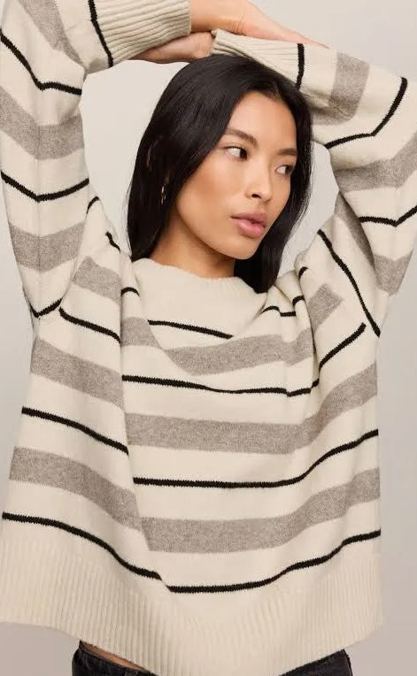A woman wearing a cream and gray striped knit sweater with black horizontal stripes, posing with her arms raised against a light gray background.