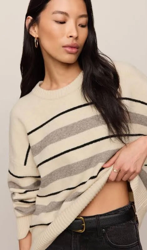 A woman wearing a cream-colored knit sweater with horizontal black and gray stripes, paired with black jeans, against a neutral beige background.