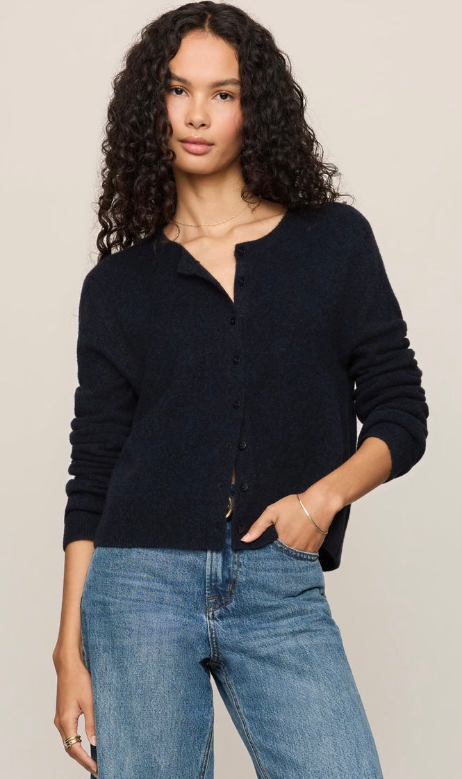 A woman with curly dark hair wearing a dark navy blue button-up cardigan and blue jeans, standing against a plain light beige background.