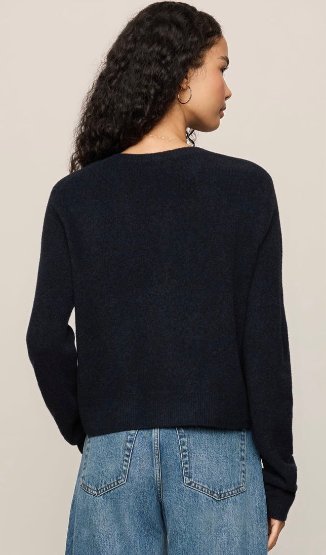A woman shown from the back wearing a dark navy knit sweater and blue jeans against a plain light beige studio background.