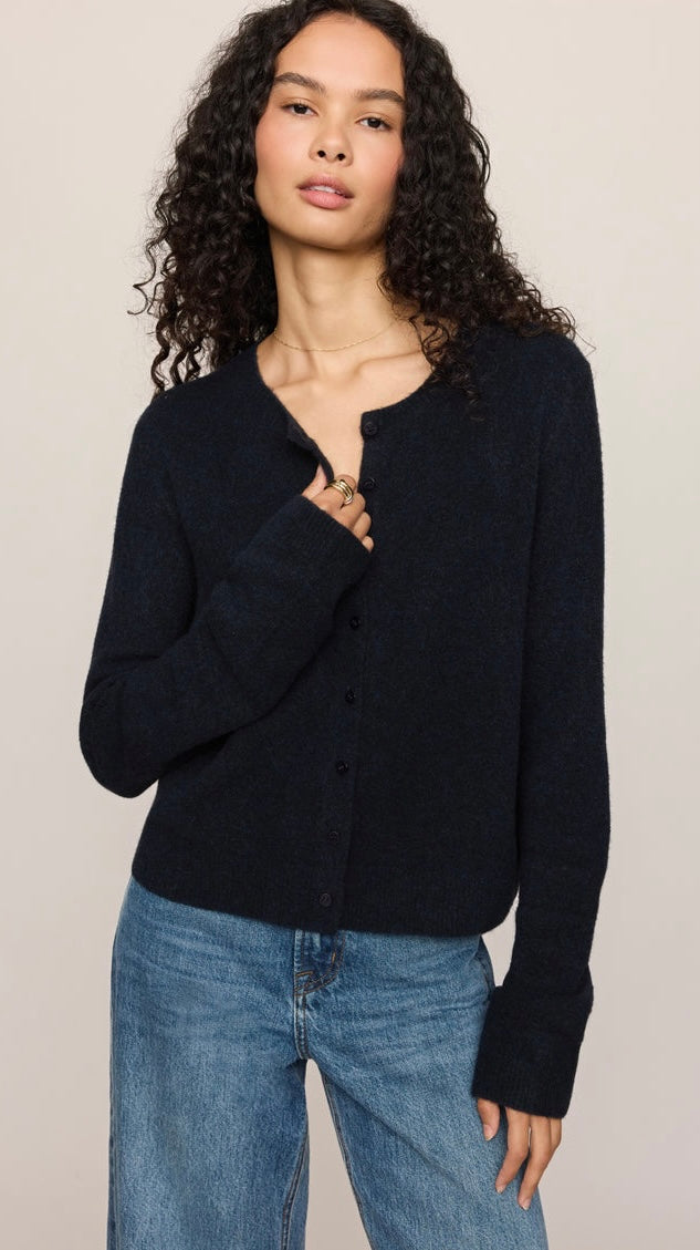 A woman with curly dark hair wearing a dark navy blue knit cardigan with a split neckline and button front, paired with blue jeans, against a plain light beige background.