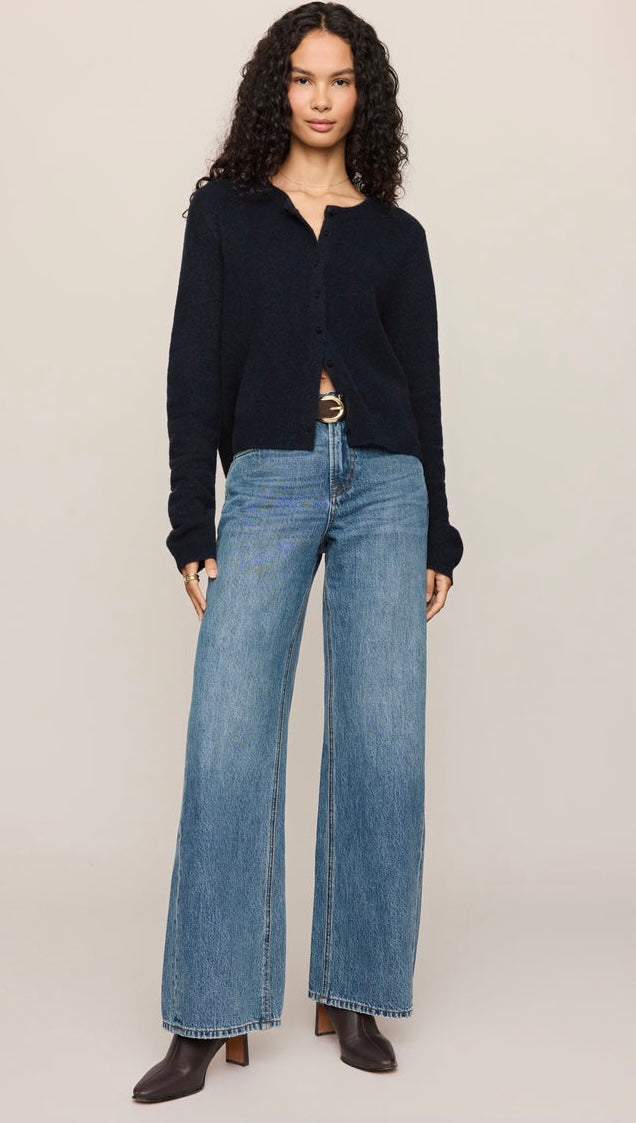 A model wearing a dark navy long-sleeve knit cardigan, wide-leg blue jeans, and dark brown heeled boots, standing against a solid light beige background.