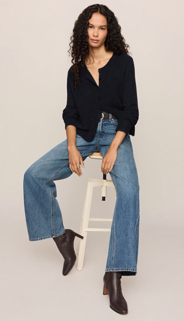 A model sits on a white stool wearing a black long-sleeve button-down shirt, wide-leg blue jeans, and dark brown ankle boots against a light gray studio background.