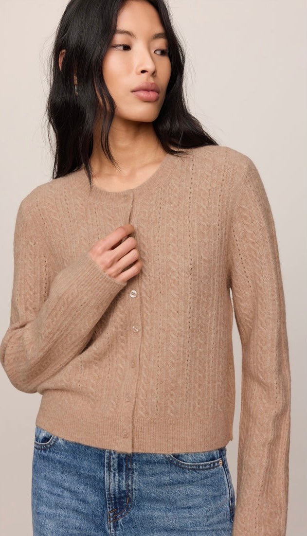 A woman wearing a cropped, long-sleeve cable-knit cardigan in a warm beige/tan color, paired with blue jeans, against a plain light beige studio background.