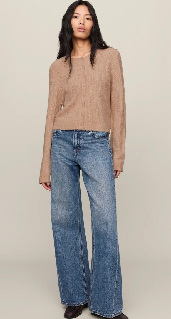 A woman wearing a cropped, long-sleeve cable-knit cardigan in a warm beige/tan color, paired with blue jeans, against a plain light beige studio background