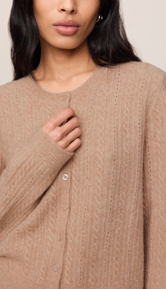 A close-up of a woman wearing a beige cable-knit button-front cardigan sweater against a plain light background.