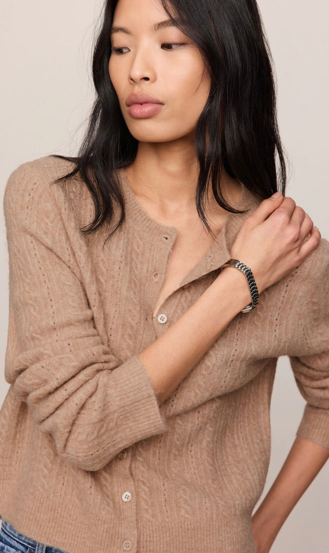A woman wearing a beige cable-knit button-up cardigan sweater, shown from the chest down against a plain light beige background.