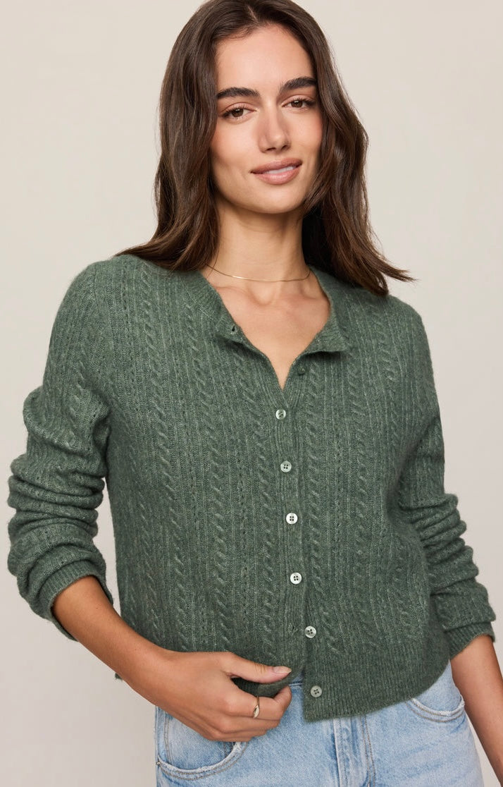 A woman wearing a sage green cable-knit button-up cardigan sweater and light blue jeans, standing against a plain beige studio background.