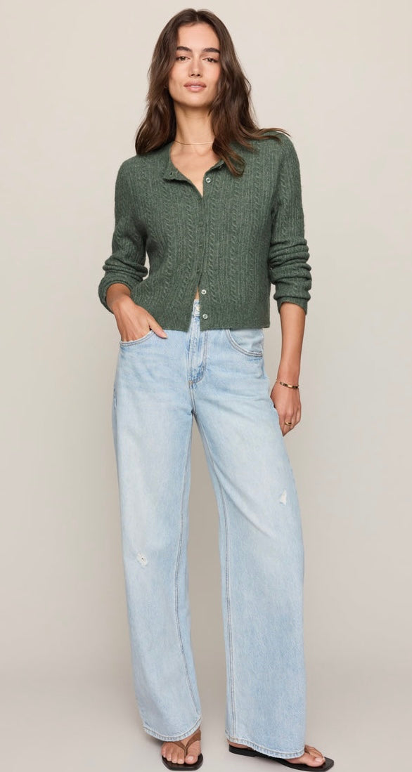 A woman modeling a dark green cable-knit cropped cardigan with light-wash wide-leg jeans and black sandals, standing against a solid light beige background.