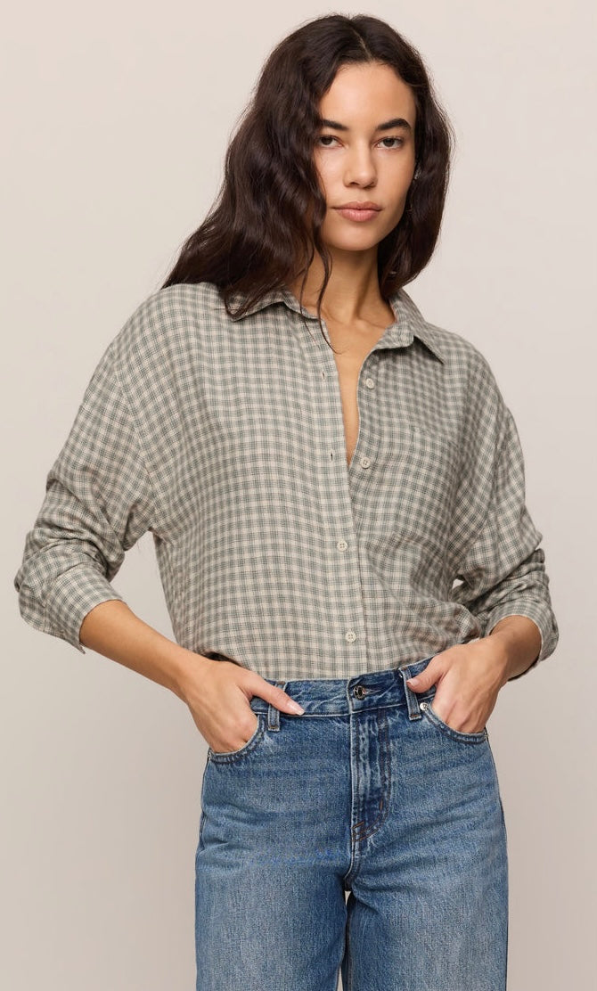 A woman wearing a muted sage green and white gingham button-down shirt tucked into blue jeans, standing against a solid light beige background.