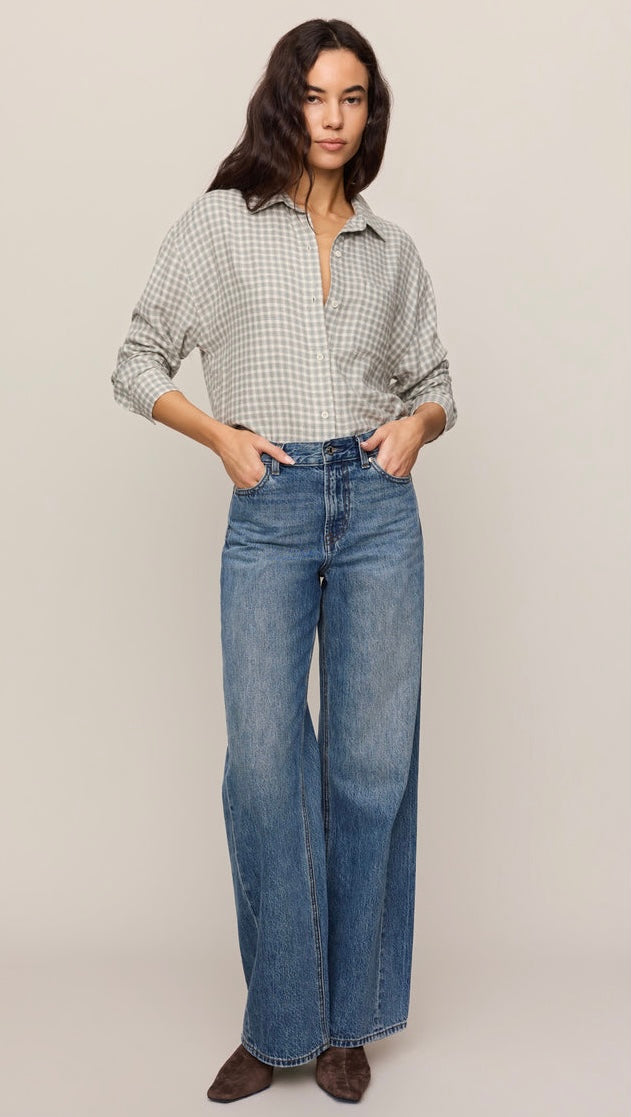 A woman modeling a light gray and white gingham button-down shirt paired with high-waisted, wide-leg blue jeans and brown suede boots, set against a plain beige studio background.