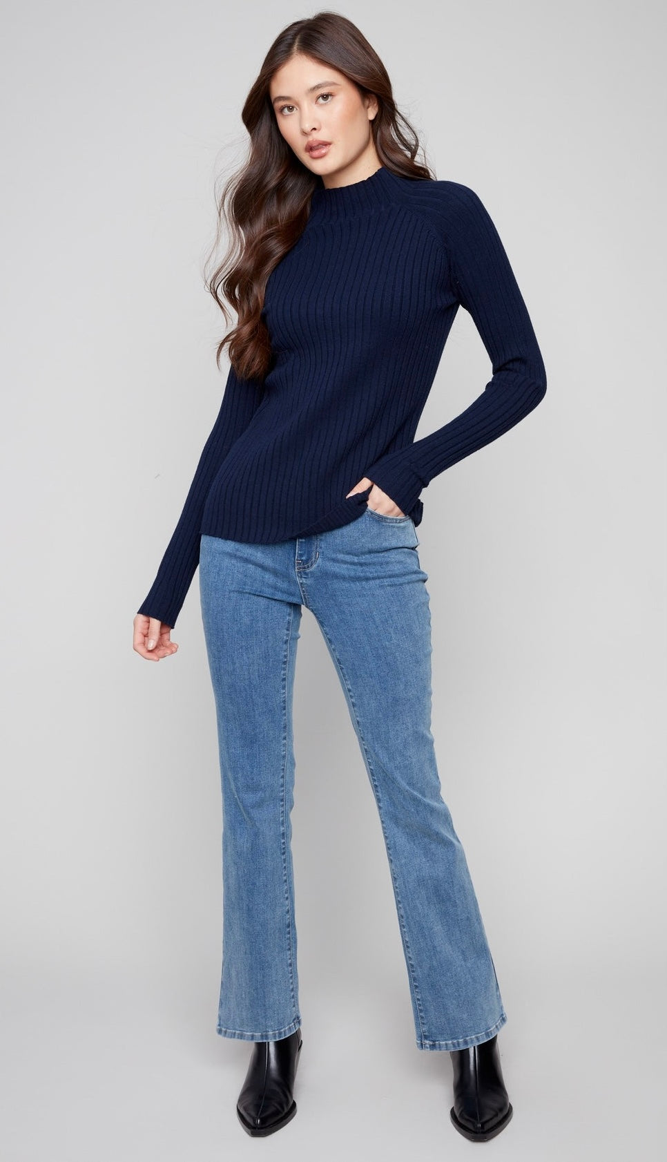 A woman modeling a dark navy ribbed mock neck sweater paired with light blue flare jeans and black ankle boots, standing against a light gray studio background.