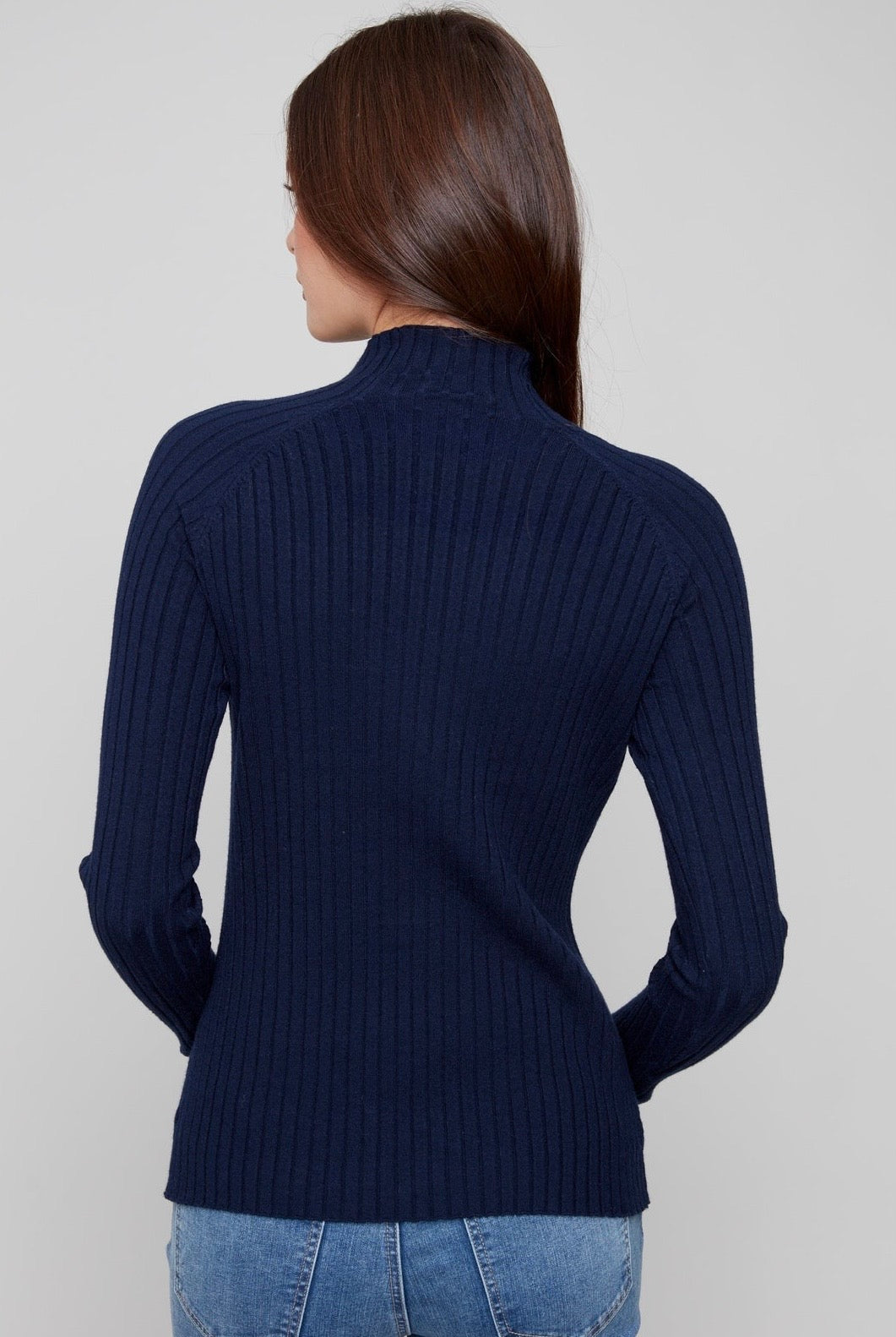 A woman viewed from the back wearing a navy blue ribbed mock-neck sweater and blue jeans against a light gray studio background.