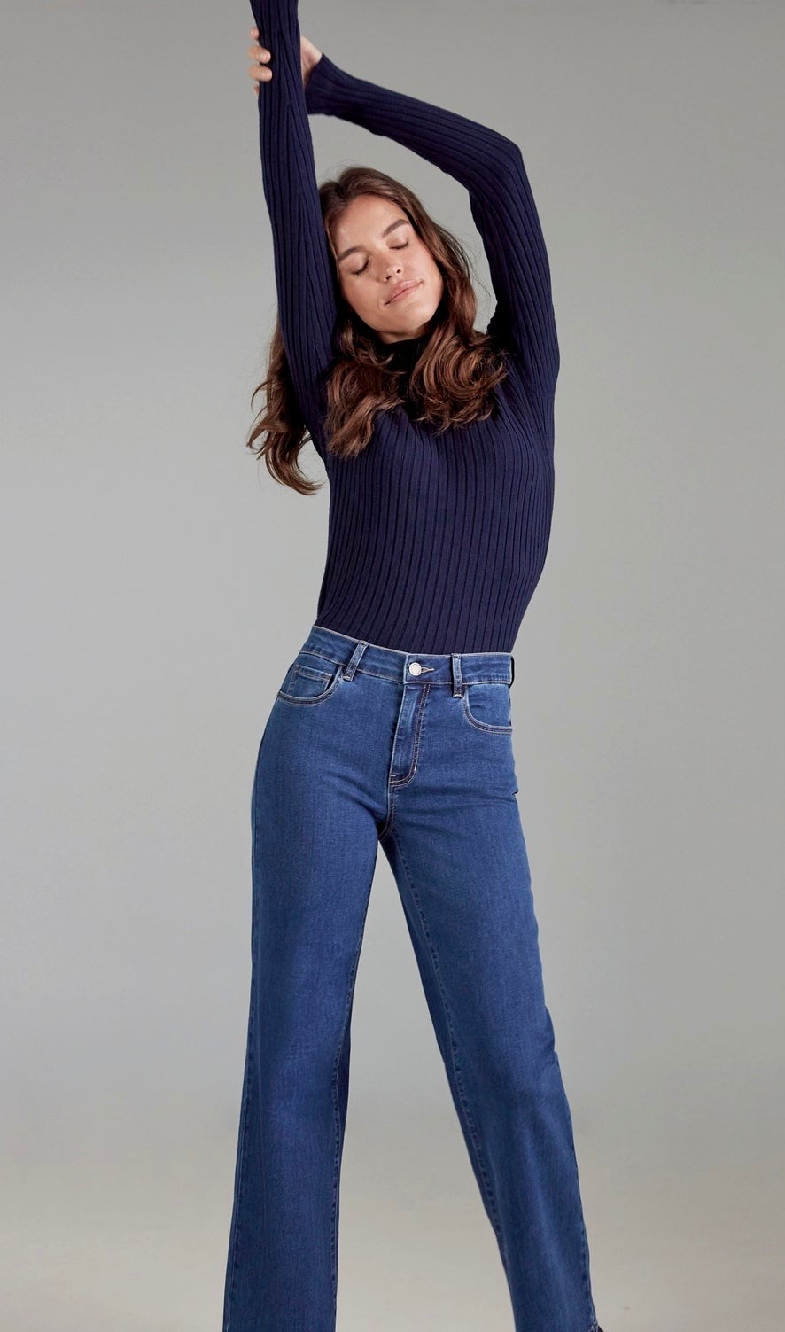 A woman modeling a dark navy ribbed long-sleeve top and blue jeans against a plain light gray studio background.