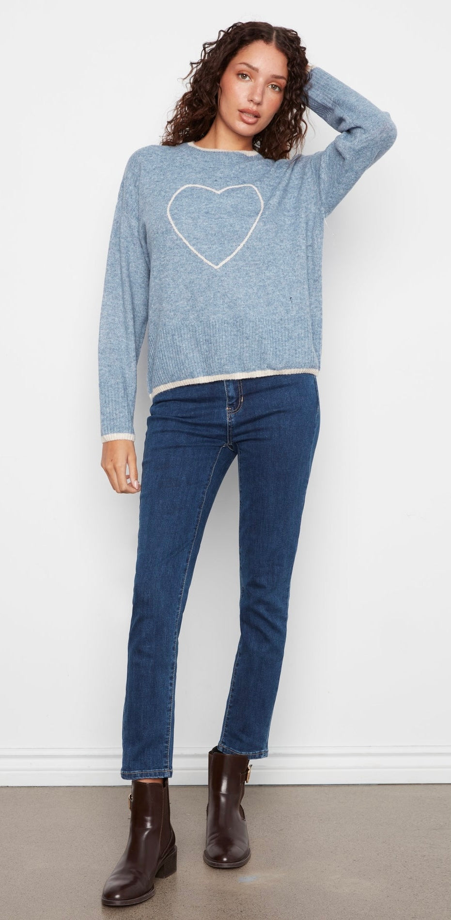 A woman modeling a light blue heathered long-sleeve sweater with a white heart outline on the chest, paired with dark blue jeans and brown ankle boots, standing against a white wall.