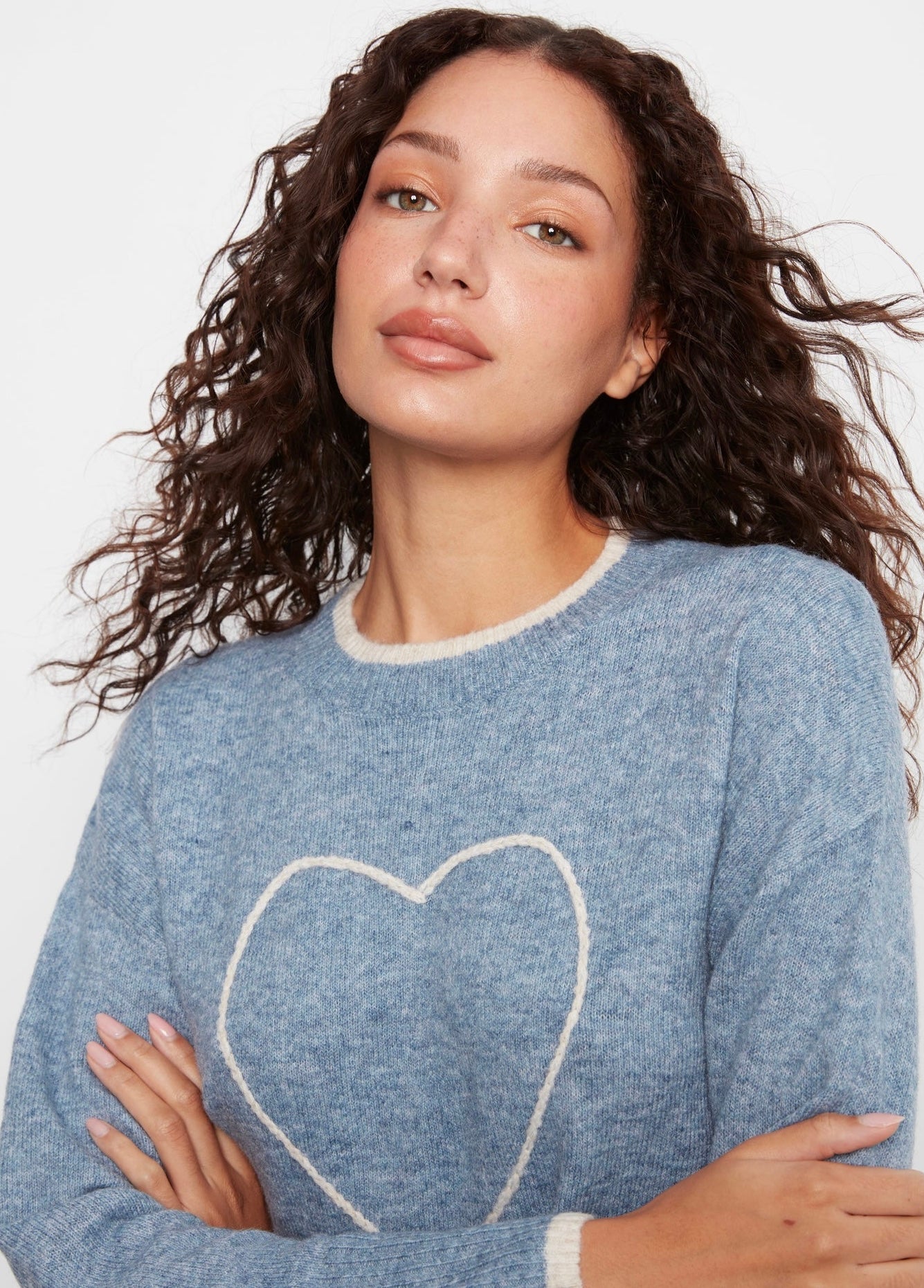 A woman with long, curly brown hair wearing a heathered blue crewneck sweater with a white knit heart outline on the front, set against a plain light gray background.
